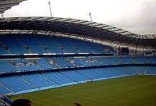 Roughly the same camera position shows grass up to the blue seats of the stands. The stand is now split into three tiers of permanent seating.