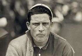 A man in a baseball jersey and a white sweater, which is worn over only his right sleeve. The man is also wearing a light-colored baseball cap with a "C" on the front. His face is freckled, and his eyes look directly into the camera.