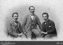 Three young men in suits with high white collars and bow ties, sitting.