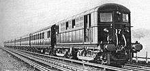 A black and white image of an electric locomotive hauling at least 6 coaches, shown with the electric locomotive on the right. A track in the foreground is electrified with the fourth rail system. The locomotive is shown with two pick-up shoes.