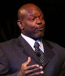 A man wearing a black striped suit and black tie with white dots.