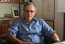 A grey-haired bespectacled man wearing a light blue shirt and sitting on a chair behind a desk; behind him is a bookshelf and a wall mounted with certificates and awards.