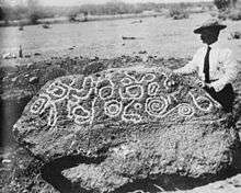 Photo taken in 1910 in Mexico of the very large Esperanza Stone, with Burnham standing to the right of the stone. The many inscriptions on the stone are sort of circular and have been filled in with white flour so they can be easily photographed.  In the background is a desert landscape.