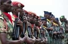 A row of uniformed men stand in formation, with guns at shoulders