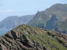 Ricky peaks and cliffs topped by greenery, with a small section of blue see visible to the left.