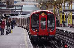 A train is slowing to stop at a platform on the right. Although there is a roof, sunlight can be seen through gaps; another platform and track can be seen on left. People are standing or walking on both platforms.