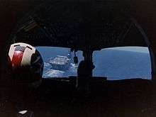 Photograph of the interior of an aircraft cockpit, taken from behind the pilot, who is sitting on the left hand side. An aircraft carrier is visible through the left-hand pane of the cockpit windows.