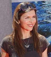 A woman with long brunette hair, wearing a grey top also with a necklace.