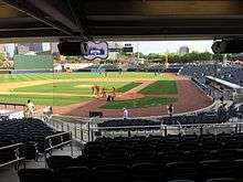 A green baseball field on a sunny late afternoon and blue seats surrounding the field viewed under the shade of the concourse