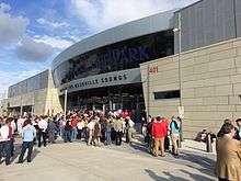 A crowd of people waiting to enter the ballpark through a concrete, metal, and glass facade on a sunny late afternoon