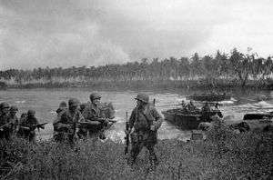 Soldiers walk through long grass. Other soldiers are arriving in landing craft in the lagoon behind them. In the background is a coconut plantation. The sky is overcast.