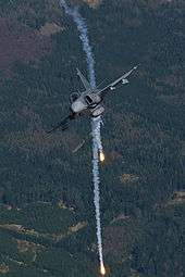 Head on view of fighter jet banking right while releasing flares against a background of green woodland.