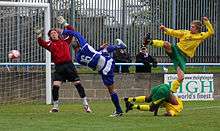 Several players standing near a goal, attempting to reach the ball. Some of them are not touching the ground, and do not appear to be completely in control of their bodies