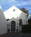 A white stuccoed hall, partly in shadow and linked at its left corner to the wall of a larger building, which partly obscures it. An entrance porch has a grey double-door and a semicircular transom light, two lamps and a plaque reading "Gospel Hall".  The porch obscures two semicircular-arched windows, above which is a pediment with a round window.