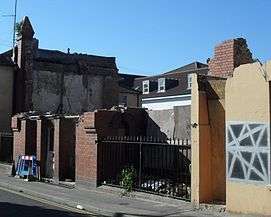 A vacant space is surrounded by a weathered wall and gate. Behind the space stands two buildings, the one on the left looking somewhat decrepit while the one in the middle looking better taken care of.