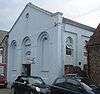 Three-quarter view of a pale blue building with a plain exterior and a large pediment, on a crowded site between two flint structures.  The façade has three tall, triple-recessed blank arches, the centre of which is shortened to accommodate an entrance door.  A round opening sits just below the pediment.  The side has round-arched windows.