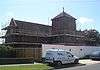 A long brick and tile building with central tower on the near side.  The whole structure is surrounded by scaffolding and barricades.  The tower has tall round-headed windows and louvres on each face, a shallow tiled cap and a stone crucifix.