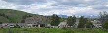 alt=Photograph of Fort Yellowstone, showing several Army buildings, modern motor vehicles, and the wide-open Yellowstone landscape in the background.