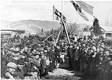 A black-and-white photograph of a large group of people wearing hats standing in front of two flagpoles flying flags, one with the letter "W" on it