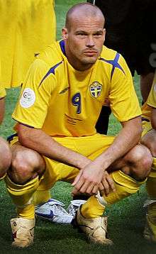 A young man with a shaven head, wearing a yellow shirt. His arms are above his head, and he appears to be clapping