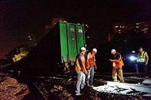 Three men wearing orange reflectorized safety vests and hard hats standing around a section of track at night looking at a portion of rail illuminated by a flashlight held by a fourth man standing behind a third rail. Next to him is another man wearing a vest but no hard hat. To the left is a large green metallic box on a flatcar