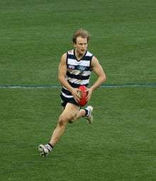 Young male athlete runs while wearing a navy blue and white striped sleeveless shirt and blue navy shorts. He looks ahead of the field as he holds the red football and prepares to kick it.