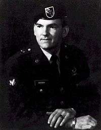 Head and torso of a white man wearing a beret with a shield-shaped patch on the front and a dark military jacket. His hands are crossed in front of him; on a finger of his left hand is a ring and on his right hand is a metal bracelet.