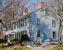 A blue colonial two story house with black shutters. Over the front door is a flag pole holding an American flag.