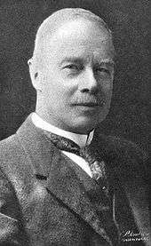 Head-and-shoulders portrait of a sixtyish man in a wingtip collar and tie and a three-piece suit. He is nearly bald and wears a mustache. He looks directly at the viewer with the vestiges of a smile.