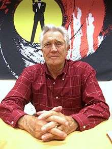 Grey haired man in red check shirt, sitting at a table on which his hands are resting.