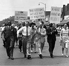 A group of four middle-aged men in suits and one woman in a dress walk in the first rank of a procession of individuals down the middle of a street. Brick upper-stories of storefronts appear in the background, from middle to the right; tops of trees appear in the distance, far left. Three placards tacked onto pickets and held by two men in the second rank and one in the first rank read as follows. "I Am John A. Maxwell I Was Discriminated Against In The Pointes." "The Freedoms We Lose May Be Your Own." "A House Holds No Prejudice."