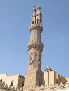 An ornate carved stone octagonal minaret, with a carved stone railing around balconies at its center and near its top. Above the second balcony the minaret splits into two rectangular shafts, each tipped by railing and a bulb-shaped finial.