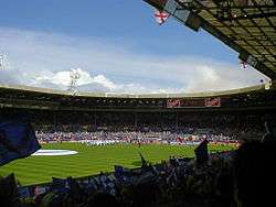 A sports stadium full of spectators.  Those nearest the camera are waving blue and white flags.