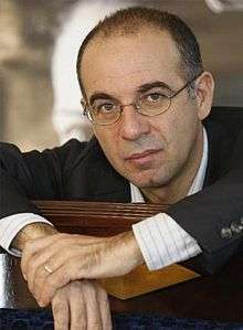 A photo of an Italian male who is seen with his hands clasped together. He is wearing glasses, a black coat, an white collared shirt with stripes