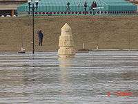 Water from a river completely surrounds a stone obelisk. In the background are several people with news cameras. A building with a green roof is seen behind them. At the bottom right of the image is the camera's time: "6 7:59 am".