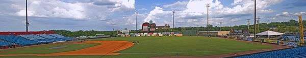 A panorama of the ballpark taken from the right field seats showing the field, entire outfield wall, guitar scoreboard, green trees outside the park, and a partly cloudy blue sky above