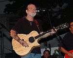 A man in his 50s or 60s, wearing a black top and jeans, with jewellry on both wrists and a crucifix around his neck. He is playing an acoustic guitar, and standing in front of a microphone. Other musicians are also visible.