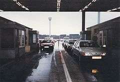 View of two lines of vehicles passing between two buildings, with four passport control booths visible, under a corrugated metal roof. A long line of vehicles stretches into the distance below towers ringed with searchlights.