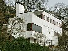 A small, flat-roofed, stucco house designed with simple, rectangular shapes, and built into a hillside. The casement, paned, and plate glass windows are wood framed.