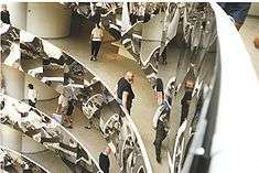 The same rotunda with the balconies covered in mirror-polished stainless steel.  There are people walking along galleries, and a burly man with large beard, wearing, a black t-shirt, leans on the railing.