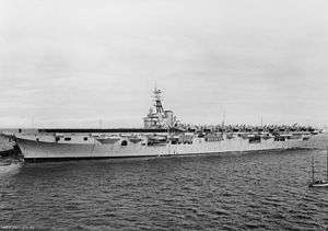 An aircraft carrier travelling at slow speed. Naval personnel in black uniforms are lined up on the forward half of the flight deck, while propeller aircraft with folded wings crowd the back half.