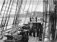 The deck of a sailing ship, viewed from slightly above. Naval cannons protrude through embrasures on this ship's side. About two dozen sailors are on deck. Standing rigging runs from the gunwales to the unseen masts overhead. A raised deck is at the rear, with a boat on davits extended outboard. The ship appears to be in a port, with hills in the background.