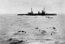 A large warship lowers boats to pick up sailors floating in the open water