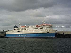 A large modern ferry with a blue hull and white topsides lies next to a harbour under grey skies.