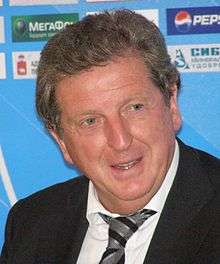 A photograph of a grey-haired, middle-aged man at a press conference. He is wearing a black suit, a white shirt and a grey and black striped tie. He is speaking with someone out of frame.