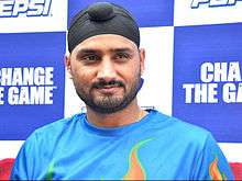 A Black-bearded man wearing a white turban stares directly at the camera. He is wearing a grey-blue and white shirt with the colours of the Flag of India and the word "SAHARA" on the shirt's chest area.