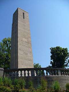 A photograph of a grey-bricked structure with green trees in the background and foliage in the foreground all under a clear, blue sky