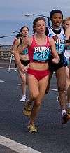 A woman in red runs alongside a group of athletes on a grey road.