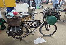 A profile of Heinz Stücke's black bicycle, laiden with baggage and a bedroll. Behind it, Stücke talks with two visitors.