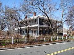 A roughly square house with porches wrapped around all sides. Its yard has trees with no leaves on them.